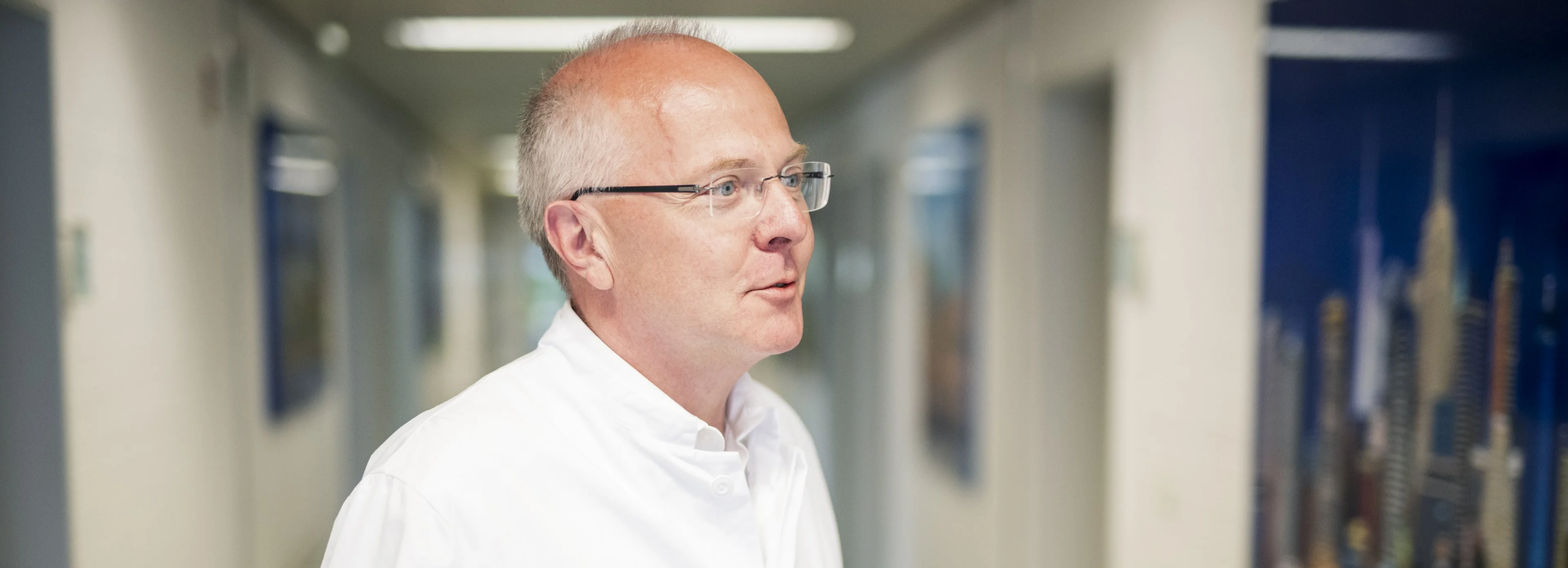 Ein Mann mit Brille und weißem Laborkittel gestikuliert mit den Händen, während er in einem Flur mit weißen Wänden und gerahmten Bildern steht.