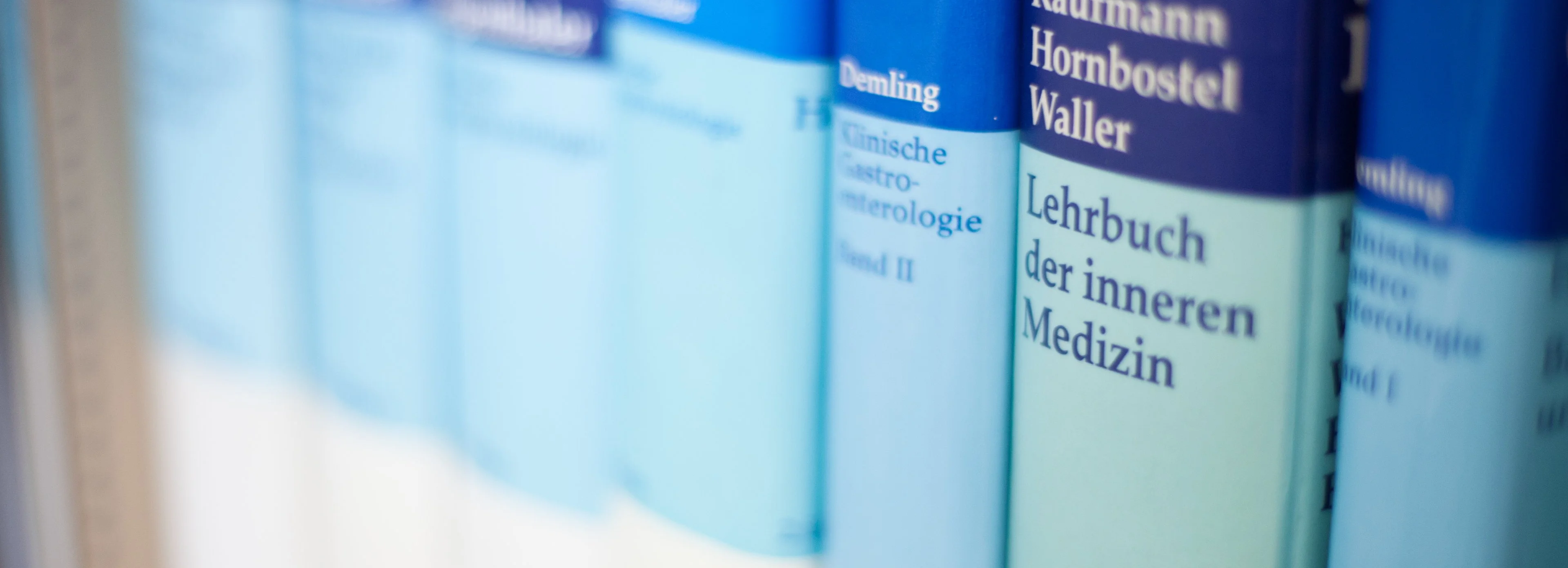 Eine Reihe blau-weißer deutscher Medizinlehrbücher in einem Regal, deren Buchrücken teilweise beschädigt sind und deren Titel erkennbar sind, darunter „Lehrbuch der inneren Medizin“.