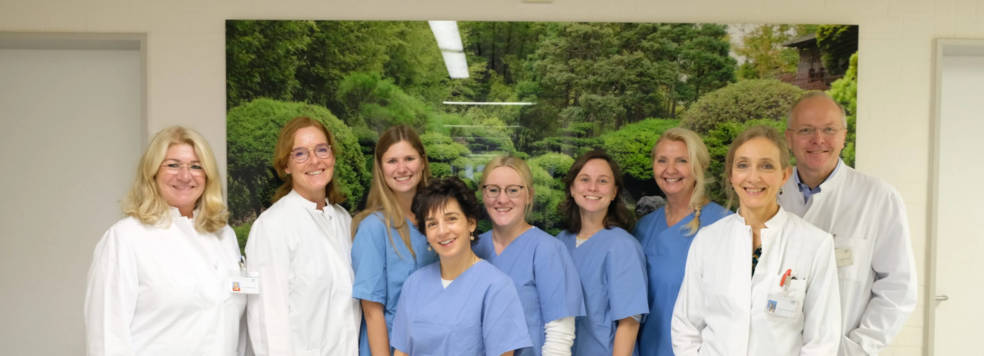 Eine Gruppe von neun medizinischen Fachkräften, von denen einige weiße Kittel und andere blaue OP-Kleidung tragen, steht gemeinsam lächelnd in einem Flur, hinter ihnen hängt ein großes Naturfoto an der Wand.